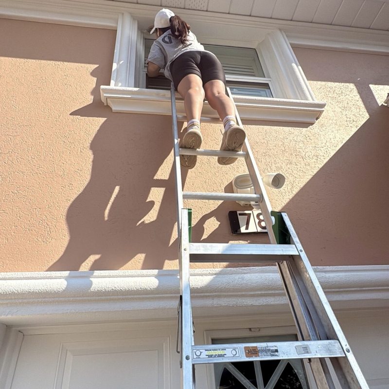 Worker painting window trim on an upper story while standing on a ladder.