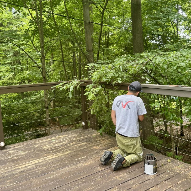 Worker kneeling while staining or painting a wooden deck surrounded by trees.