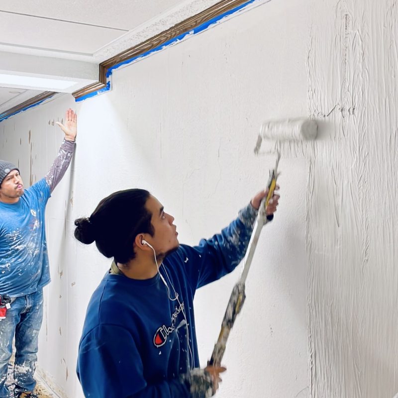 Painters applying white paint on an interior wall using rollers and extension poles.