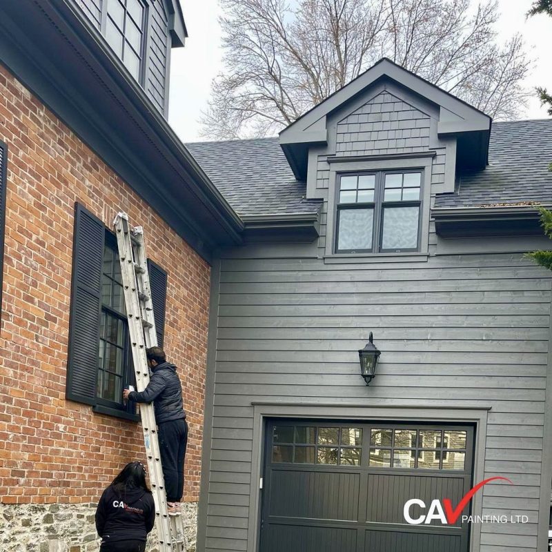 Two painters working on exterior window frames of a gray and brick house.
