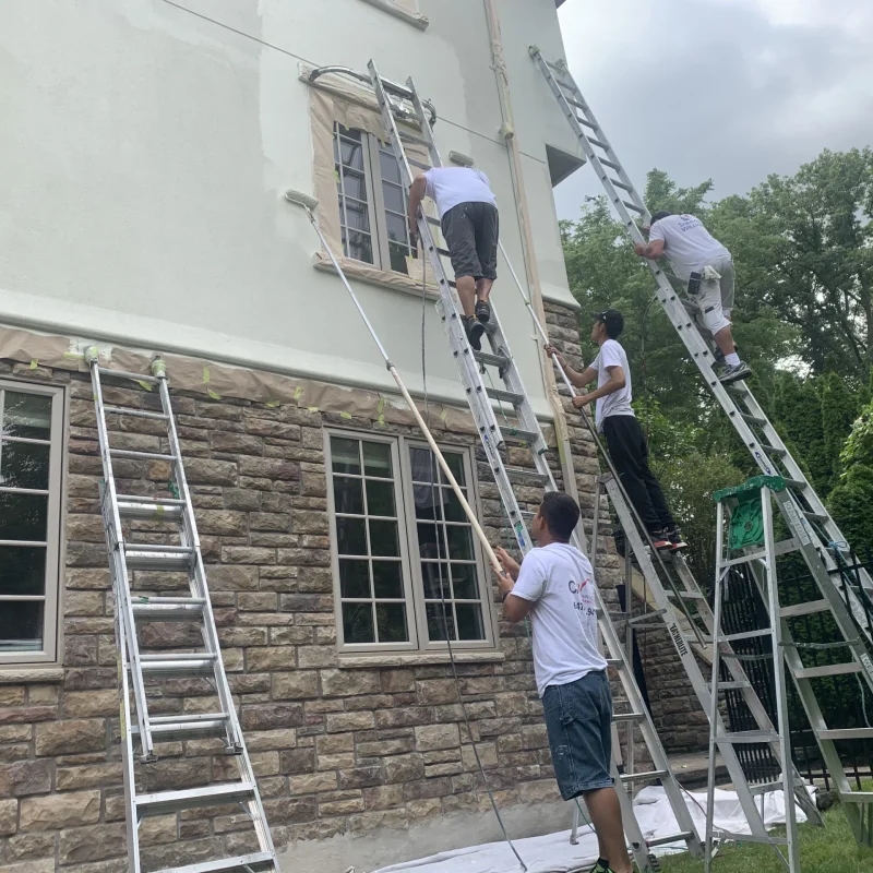 Group of painters applying paint to a house facade using multiple ladders.