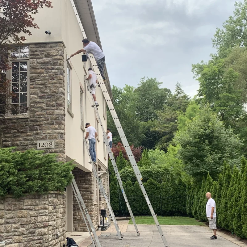 Team of painters working on a tall exterior wall using secured extension ladders.