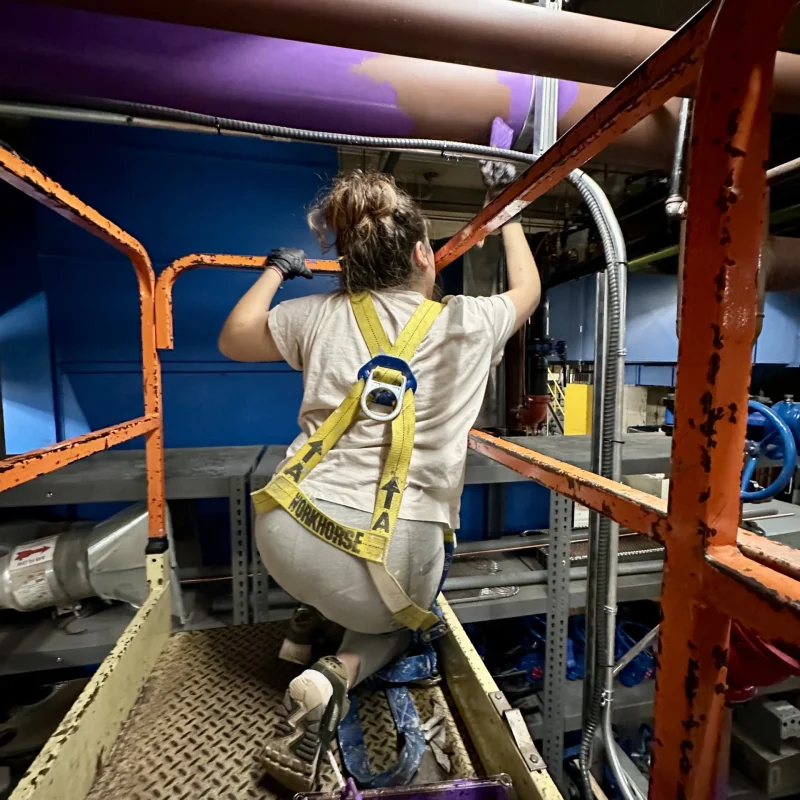 Worker painting overhead pipes on a platform while wearing a safety harness.