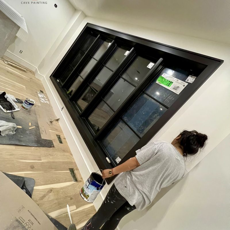 Painter applying white paint around black-trimmed windows in a modern room.
