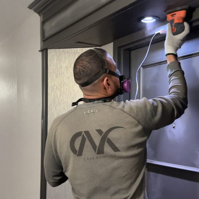 Painter sanding or prepping a metal doorway surface while wearing a respirator.