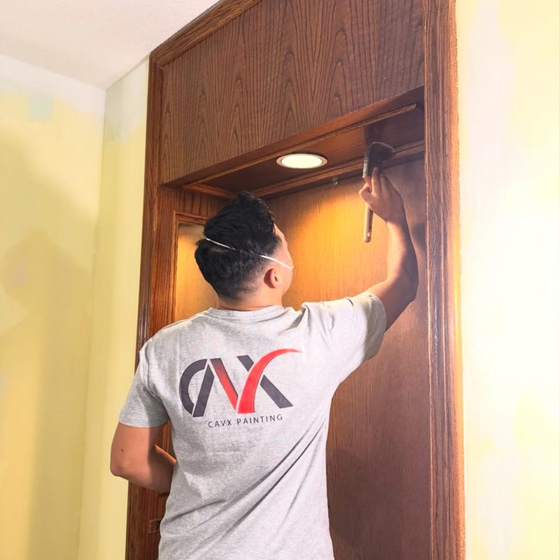 Painter brushing wood trim inside an interior doorway under focused lighting.