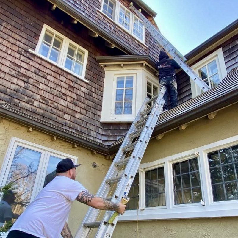 Two painters setting up ladders to reach upper windows on a multi-story home.