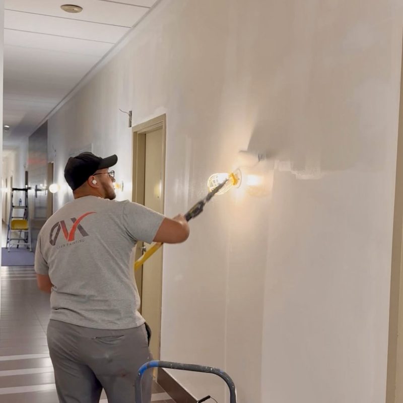 Worker applying beige paint to a hallway wall using an extension roller.