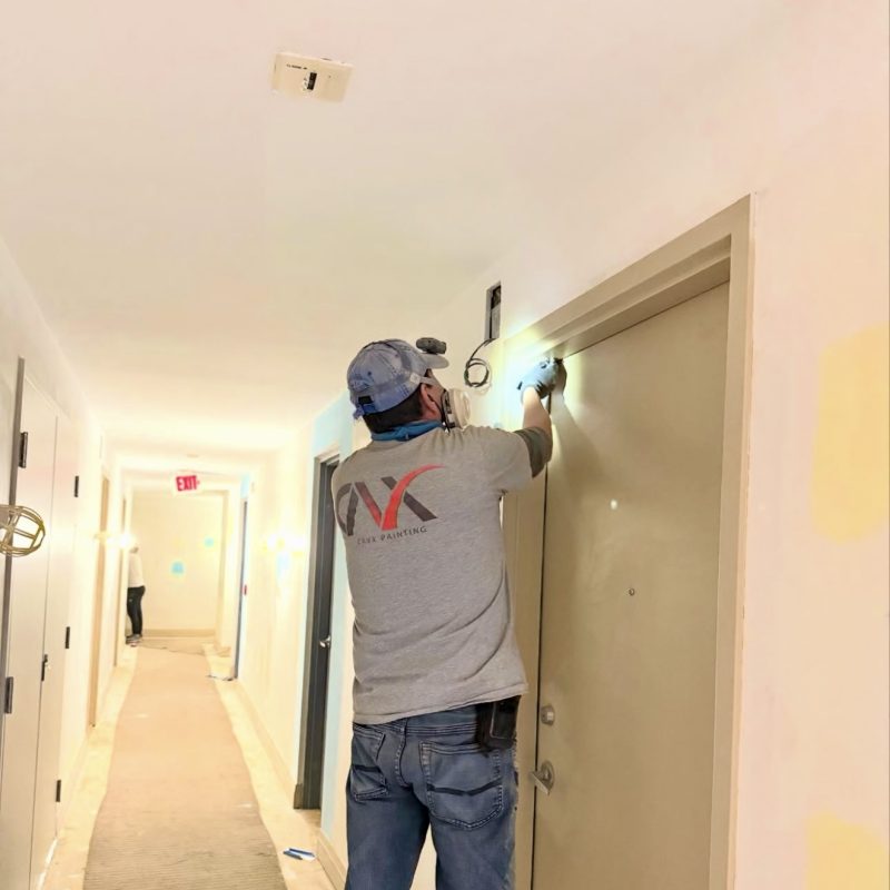 Worker painting a door frame in a brightly lit apartment hallway.