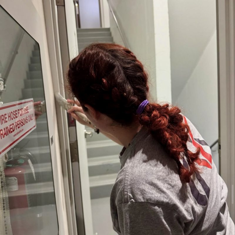 Worker painting a door frame near a stairwell while wearing a gray shirt.