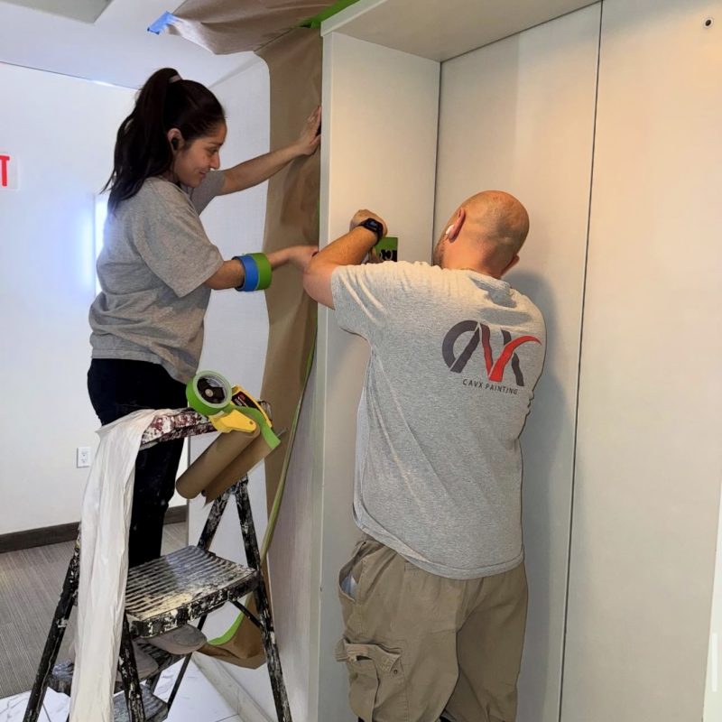 Two painters taping and preparing walls for painting in a well-lit hallway.