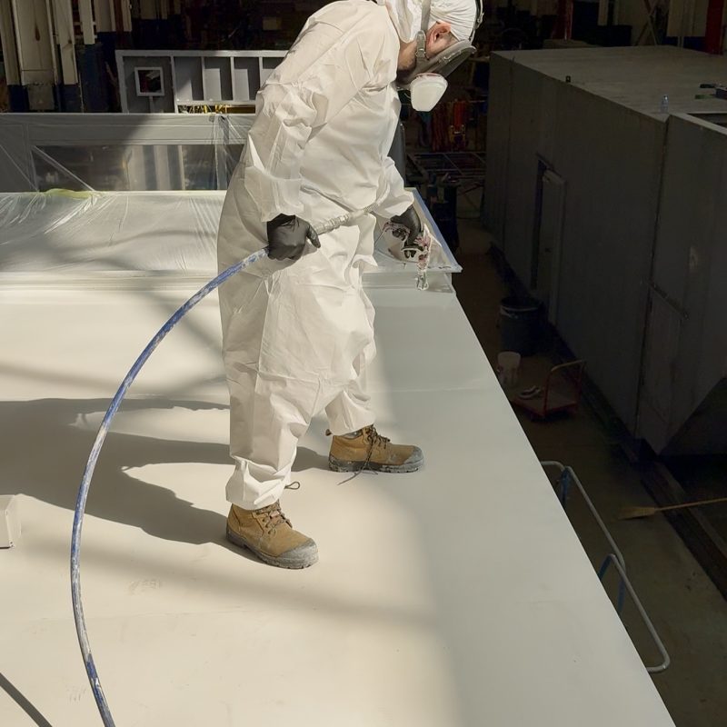 Worker in protective suit spraying paint on a large industrial surface.