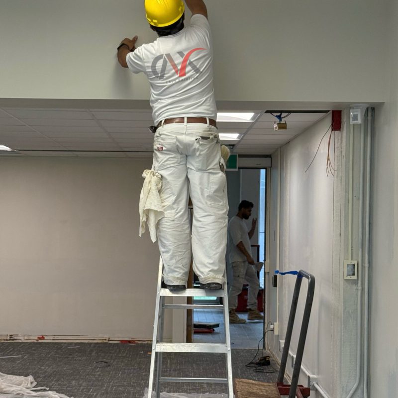 Worker on a ladder painting near ceiling lights wearing a yellow hard hat.