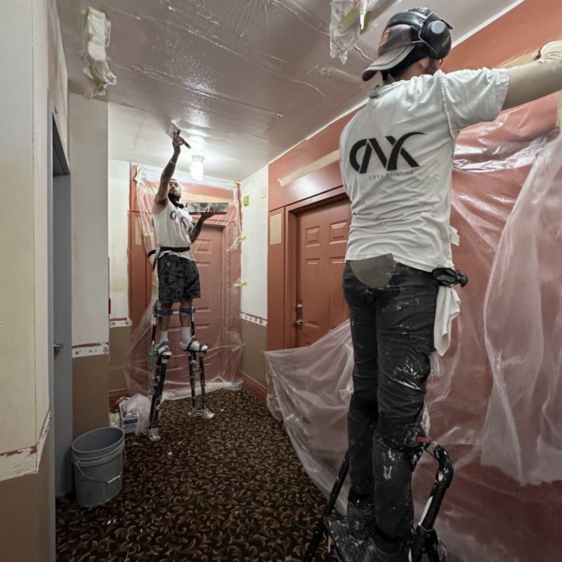 Painters using stilts and plastic coverings while painting a hallway ceiling.