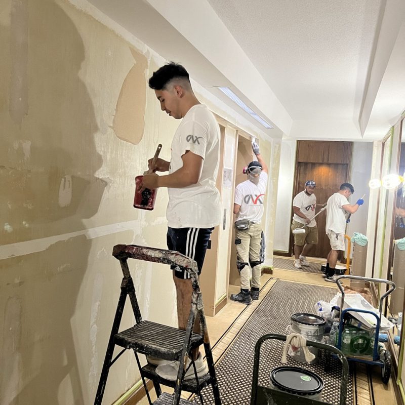 Team of painters prepping and painting hallway walls with ladders and tools.