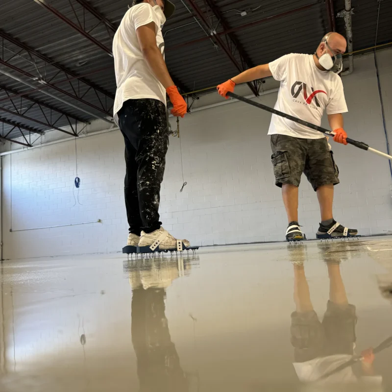 Two workers applying epoxy coating on a concrete floor using rollers.