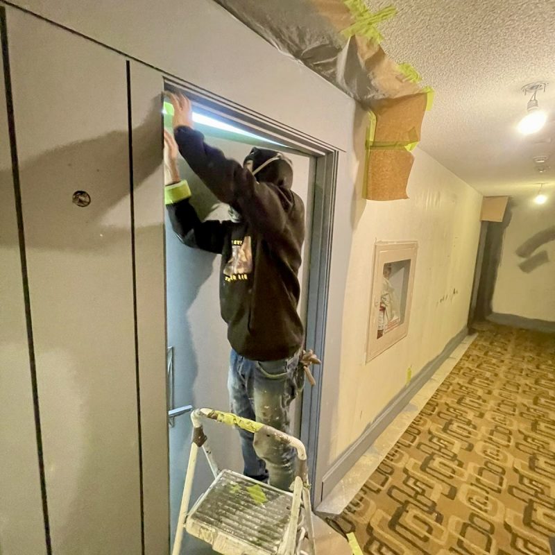 Worker taping and prepping a doorway for painting in a building hallway.