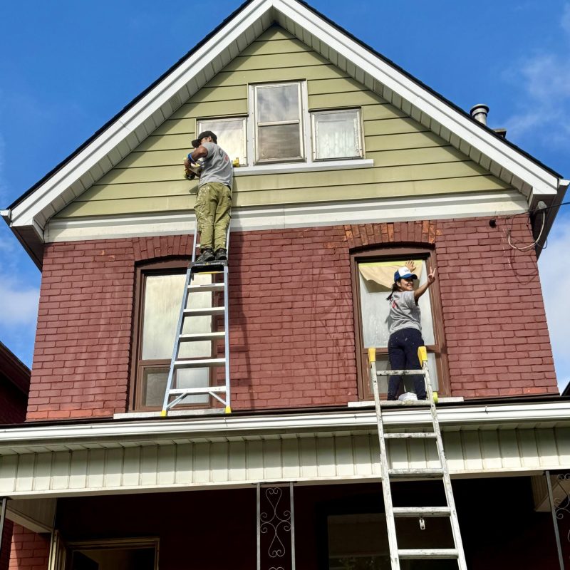 Two painters on ladders repainting the exterior of a red brick residential house.