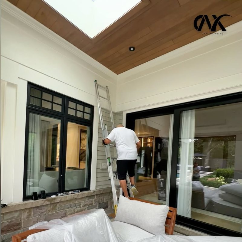 Painter on a ladder working on high interior walls under a wood ceiling.