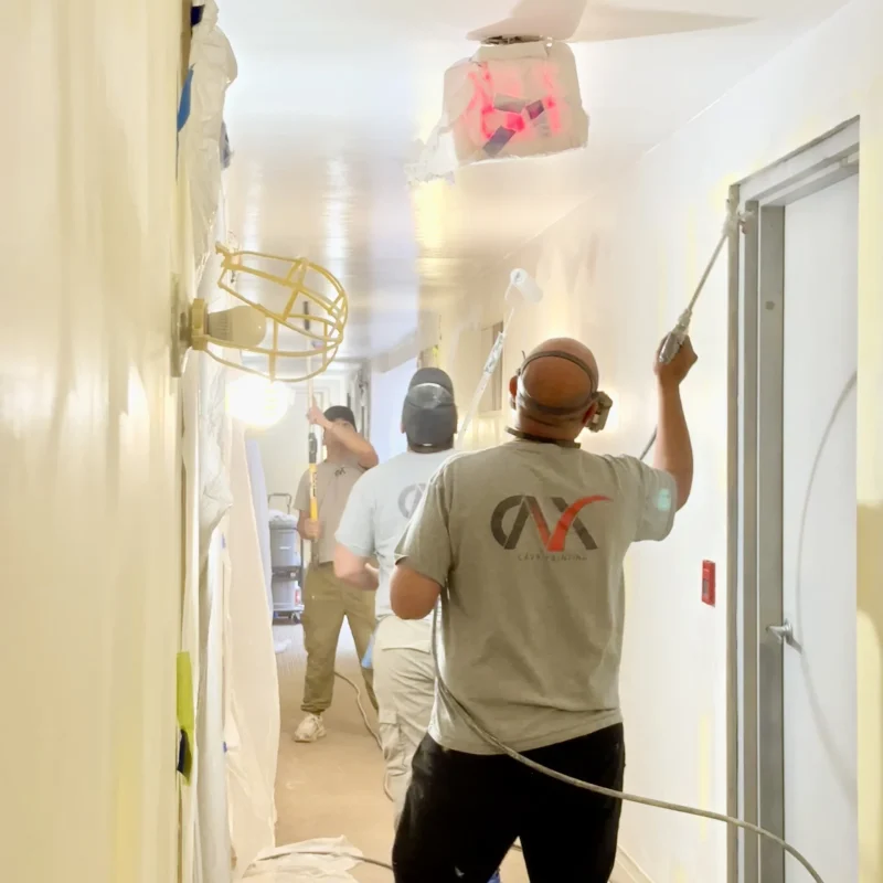 Painters spraying white paint on a hallway ceiling with protective coverings.