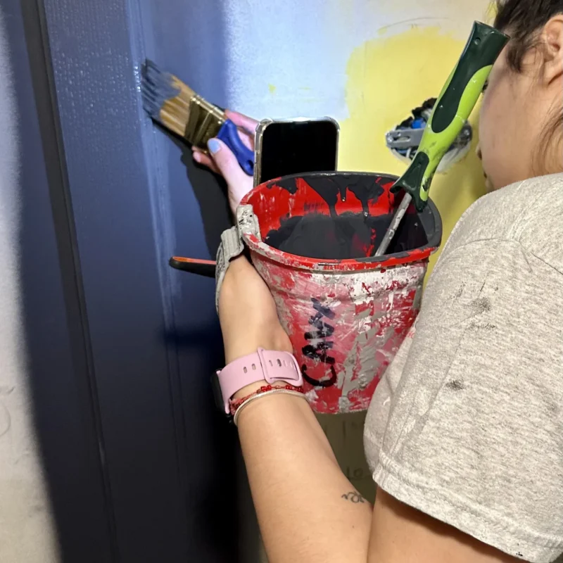 Worker painting a dark-colored door frame with a brush and red paint bucket.