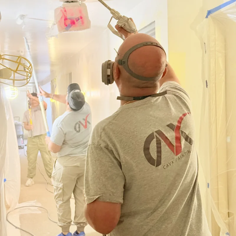 Painters spraying ceiling paint in a covered hallway using protective gear.